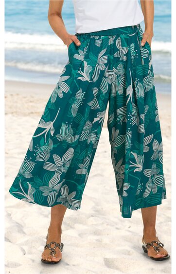 Weite, knöchellange Hose in Petrol mit großem weißen Blumenmuster, elastischer Bund, seitliche Eingrifftaschen, getragen von stehender Person am Strand