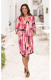 Knielanges Kleid mit auffälligem abstraktem Muster in Rosa, Pink, Weiß und Schwarz, V-Ausschnitt mit Schlitz und Bindeband, 3/4-Ärmel, gerade geschnitten, getragen von lächelnder Frau im Stehen mit Händen in den Taschen