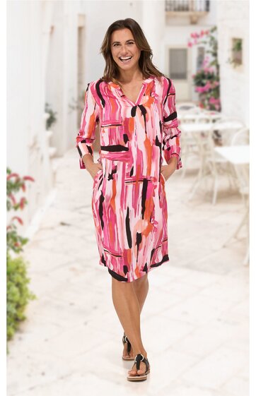 Knielanges Kleid mit auffälligem abstraktem Muster in Rosa, Pink, Weiß und Schwarz, V-Ausschnitt mit Schlitz und Bindeband, 3/4-Ärmel, gerade geschnitten, getragen von lächelnder Frau im Stehen mit Händen in den Taschen