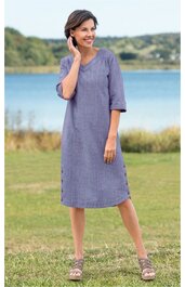 Blaues, knielanges Kleid mit kurzem, weitem Arm, rundem Ausschnitt und leichter A-Form, getragen von stehender Frau auf grünem Gras vor Wasser und Bäumen