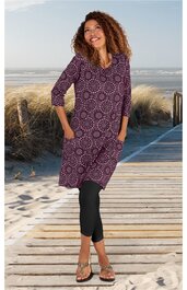 Knielanges Kleid in Dunkelviolett mit rundem weißen Kreis-Muster, U-Boot-Ausschnitt, dreiviertellangen Ärmeln, seitlichen Eingrifftaschen, getragen von stehender Frau am Strand, lockere Passform