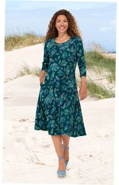 Knielanges, dunkelgrünes Kleid mit blau-grünem Blattmuster, 3/4-Ärmel, Rundhalsausschnitt, zwei seitliche Eingrifftaschen, getragen von stehender Frau am Strand