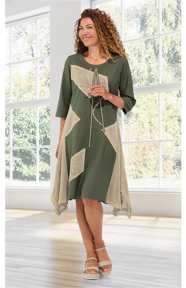 Kleid in Olivegrün mit beigen geometrischen Flächen, asymmetrischer Saum, dreiviertelärmlig, Rundhalsausschnitt mit Kordel und Quasten, getragen von stehender Frau in Innenraum vor großen Fenstern