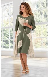 Kleid in Olivegrün mit beigen geometrischen Flächen, asymmetrischer Saum, dreiviertelärmlig, Rundhalsausschnitt mit Kordel und Quasten, getragen von stehender Frau in Innenraum vor großen Fenstern