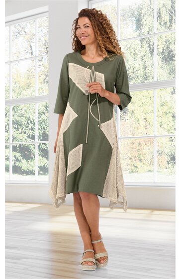 Kleid in Olivegrün mit beigen geometrischen Flächen, asymmetrischer Saum, dreiviertelärmlig, Rundhalsausschnitt mit Kordel und Quasten, getragen von stehender Frau in Innenraum vor großen Fenstern