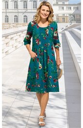 Kleid in petrolgrün mit mehrfarbigem Blumenmuster, Rundhalsausschnitt, dreiviertel lange Ärmel, Knielänge, ausgestellte Form, aufgesetzte Taschen, getragen von stehender Frau