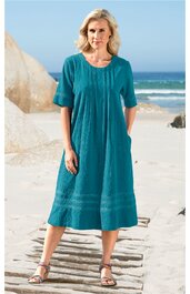 Türkiskleidschnitt knielang mit Falten vorne, Rundhalsausschnitt, halblange Ärmel mit Bund, breite Stickerei am Saum, getragen von stehender Frau am Strand, locker fallend