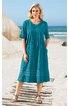 Türkiskleidschnitt knielang mit Falten vorne, Rundhalsausschnitt, halblange Ärmel mit Bund, breite Stickerei am Saum, getragen von stehender Frau am Strand, locker fallend