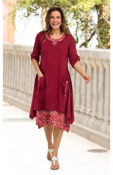 Rotes Kleid mit floral gemustertem unteren Teil, V-Ausschnitt mit Stickerei, dreiviertellange Ärmel, zwei aufgesetzte Taschen vorne, geschwungenes, asymmetrisches Saumdesign, getragen von lächelnder stehender Frau