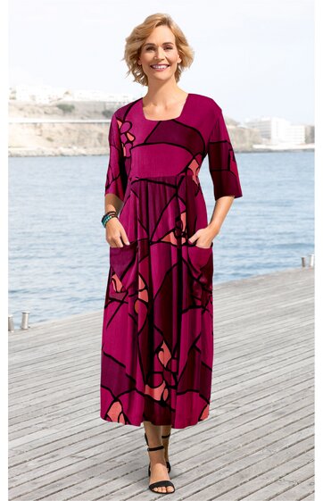 Fuchsiafarbenes Maxikleid mit abstraktem geometrischem Muster in Schwarz und Rosa, Rundhalsausschnitt, halblange Ärmel, seitliche Eingrifftaschen, fließender gerader Schnitt, getragen von lächelnder Frau stehend auf Holzsteg am Wasser