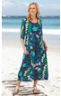 Maxikleid in Dunkelblau mit großflächigem floralen Muster in Blau, Grün und Weiß, Rundhalsausschnitt, 3/4-Ärmel, lockere Passform, ausgestellter Rock, getragen von lächelnder stehender Frau am Strand, mit Taschen