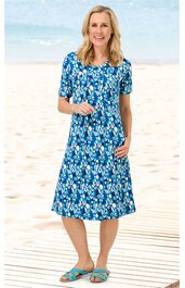 Knieumspielendes Freizeitkleid in Blau mit weißem und hellblauem floralen Muster, rundem Ausschnitt, kurzen Ärmeln, leichte A-Linie, getragen von stehender Frau am Strand