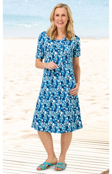 Knieumspielendes Freizeitkleid in Blau mit weißem und hellblauem floralen Muster, rundem Ausschnitt, kurzen Ärmeln, leichte A-Linie, getragen von stehender Frau am Strand