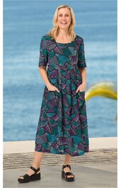 Knöchellanges Kleid in Petrol und Violett mit geometrischem Muster, rundem Ausschnitt, halblangen Ärmeln, tailliert, seitliche Eingrifftaschen, getragen von stehender Frau vor Meereshintergrund, schwarze Sandalen