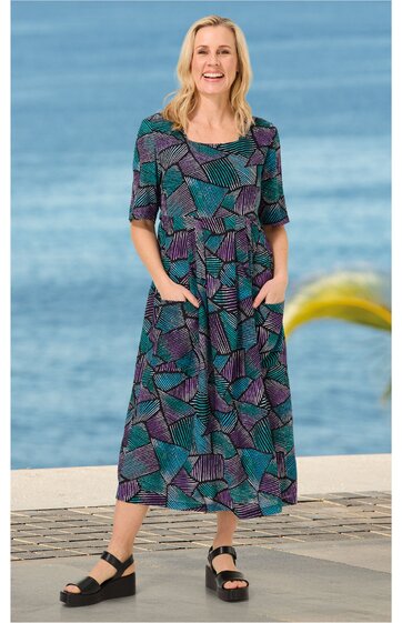 Knöchellanges Kleid in Petrol und Violett mit geometrischem Muster, rundem Ausschnitt, halblangen Ärmeln, tailliert, seitliche Eingrifftaschen, getragen von stehender Frau vor Meereshintergrund, schwarze Sandalen
