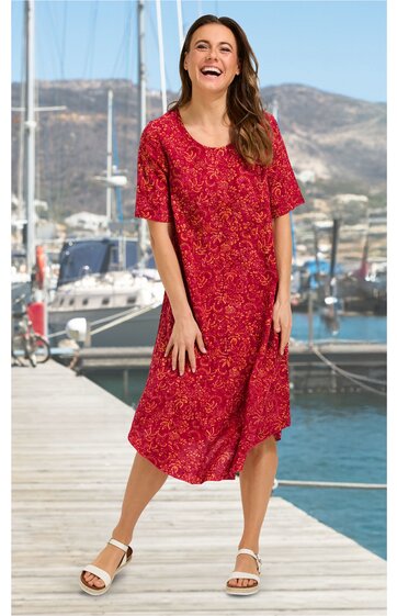 Rotes Midikleid mit floralen Muster, kurze Ärmel, runder Ausschnitt, lockere Passform, vorne rechts langer Schlitz, getragen von lächelnder stehender Frau mit offenen Haaren, weiße Sandalen