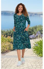 Dunkelgrünes Kleid mit floralen hellgrünen und blauen Mustern, rundem Halsausschnitt, 3/4-Ärmel, knielang, seitliche Taschen, getragen von lächelnder Frau, Stehpose, weiße Sneaker