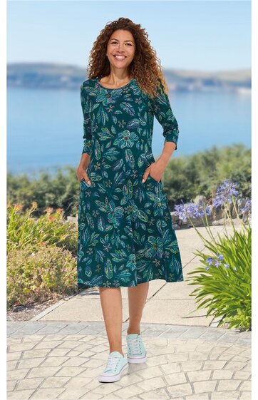 Dunkelgrünes Kleid mit floralen hellgrünen und blauen Mustern, rundem Halsausschnitt, 3/4-Ärmel, knielang, seitliche Taschen, getragen von lächelnder Frau, Stehpose, weiße Sneaker