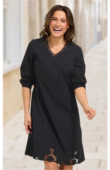 Schwarzes Kleid ohne Muster, V-Ausschnitt, lange Ärmel mit aufgekrempelten Bündchen, knielang, leicht ausgestellte Form, getragen von lachender stehender Frau