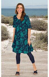 Knielanges Kleid in dunkelgrün mit türkisfarbenem Blumenmuster, Rundhalsausschnitt, halblange Ärmel, ausgestellte Form, getragen von lächelnder stehender Frau, kombiniert mit dunklen Leggings und Sandalen