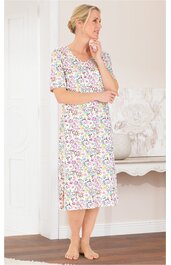 Kleid in Weiß mit buntem Blumendesign, rundem Ausschnitt, halblange Ärmel, knielang, Seitenschlitze, getragen von stehender Frau mit hochgestecktem blonden Haar