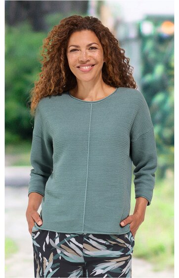 Langärmeliges, mintgrünes Damenoberteil mit feiner Struktur, Rundhalsausschnitt, lockere Passform, durchgehende Mittelnaht vorne, getragen von lächelnder stehender Frau mit Händen in den Taschen