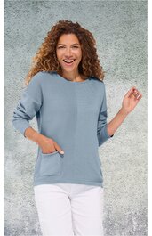 Hellblauer Pullover mit rundem Ausschnitt, lange Ärmel, aufgesetzte Tasche vorne rechts, getragen von lächelnder Frau mit lockigem Haar, weiße Hose, stehende Pose