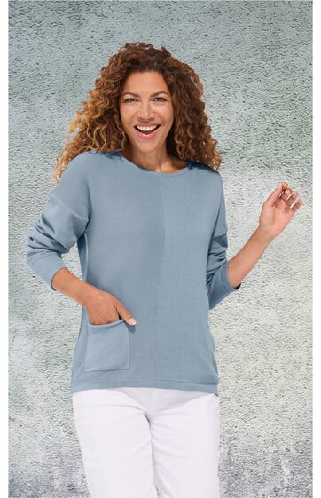 Hellblauer Pullover mit rundem Ausschnitt, lange Ärmel, aufgesetzte Tasche vorne rechts, getragen von lächelnder Frau mit lockigem Haar, weiße Hose, stehende Pose