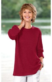 Langes, bordeauxrotes Pulloveroberteil, rundhals, lange Ärmel, aus locker fallendem Stoff, getragen von lächelnder Frau, stehende Pose, linker Arm hebt Hand zum Nacken, rechter Arm locker an der Seite