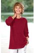 Langes, bordeauxrotes Pulloveroberteil, rundhals, lange Ärmel, aus locker fallendem Stoff, getragen von lächelnder Frau, stehende Pose, linker Arm hebt Hand zum Nacken, rechter Arm locker an der Seite