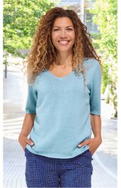 Hellblauer Pullover mit V-Ausschnitt, halblangen Ärmeln, gerade geschnittener Saum, getragen von lächelnder stehender Frau, Hände locker in dunkelblauer Hose mit kleinem weißen Muster in den Taschen