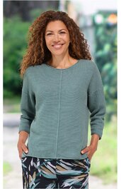 Langärmeliges, mintgrünes Damenoberteil mit feiner Struktur, Rundhalsausschnitt, lockere Passform, durchgehende Mittelnaht vorne, getragen von lächelnder stehender Frau mit Händen in den Taschen