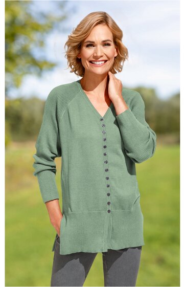 Langer, hellgrüner Strickcardigan, V-Ausschnitt, durchgehende Knopfleiste mit kleinen Knöpfen, zwei Taschen vorne, getragen von lächelnder Frau mit kurzen blonden Haaren im Freien
