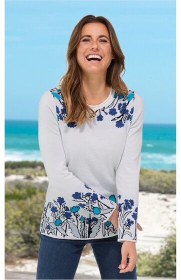 Weißer Langarmpullover mit blauem Blumenmuster auf Schultern und Saum, Rundhalsausschnitt, gerade geschnitten, getragen von lächelnder stehender Frau vor Strandkulisse