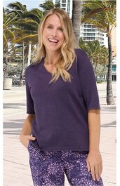 Dunkellila Kurzarmshirt ohne Muster, Rundhalsausschnitt, gerade geschnitten, getragen von stehender lächelnder Frau mit langen blonden Haaren, rechte Hand in Hosentasche