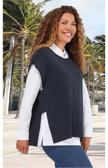 Dunkelblaue, ärmellose Weste mit seitlichen Schlitzen, rundem Ausschnitt, über weißem Langarmshirt getragen, getragen von lächelnder stehender Frau mit lockigem Haar vor Palmenkulisse