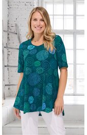 Türkisgrünes Longshirt mit abstrakten kreisförmigen Mustern in verschiedenen Blautönen, Rundhals, halblange Ärmel, ausgestellte Form, getragen von stehender Frau vor weißem Fenster und Ziegelwand