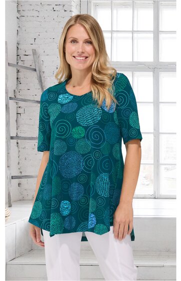 Türkisgrünes Longshirt mit abstrakten kreisförmigen Mustern in verschiedenen Blautönen, Rundhals, halblange Ärmel, ausgestellte Form, getragen von stehender Frau vor weißem Fenster und Ziegelwand