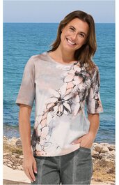 Kurzärmliges Damen-T-Shirt in beige und hellgrau mit floralem Muster in Rosa und Schwarz, rundem Ausschnitt, locker geschnitten, getragen von lächelnder Frau mit Hand in Hosentasche vor Meereskulisse