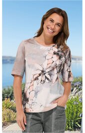 Kurzärmeliges Freizeitshirt in hellgrau mit rosa und schwarzem floralem Muster, Rundhalsausschnitt, gerade geschnitten, getragen von stehender lächelnder Frau mit Händen in den Hosentaschen vor Wasser- und Pflanzenkulisse