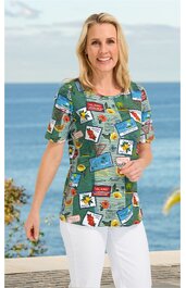 Kurzärmeliges Damenoberteil in Blau mit buntem Patchworkmuster aus Schriftzügen und Blüten, rundem Ausschnitt, gerade Form, getragen von stehender Frau vor Meereskulisse