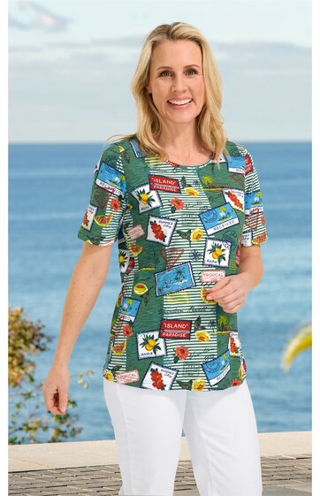 Kurzärmeliges Damenoberteil in Blau mit buntem Patchworkmuster aus Schriftzügen und Blüten, rundem Ausschnitt, gerade Form, getragen von stehender Frau vor Meereskulisse