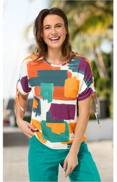 Kurzärmliges Damenoberteil mit rundem Ausschnitt, buntes geometrisches Muster in Orange, Türkis, Lila und Weiß, Ärmel mit Knoten, getragen von lachender Frau in stehender Pose