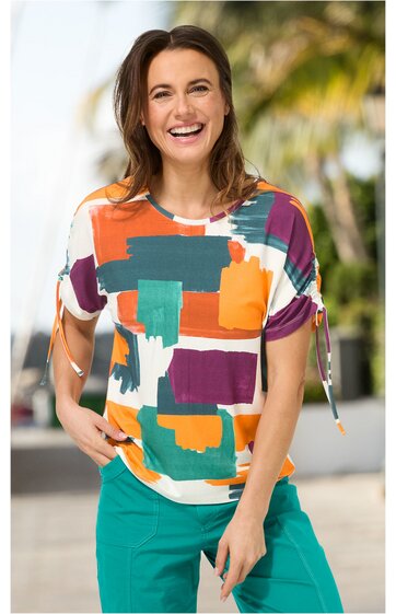 Kurzärmliges Damenoberteil mit rundem Ausschnitt, buntes geometrisches Muster in Orange, Türkis, Lila und Weiß, Ärmel mit Knoten, getragen von lachender Frau in stehender Pose