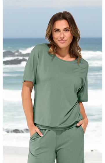 Locker sitzendes T-Shirt in salbeigrün, rundhals, kurze Ärmel, getragen von stehender Frau mit Händen in den Taschen, Strand und Meer im Hintergrund