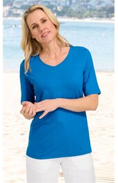 Taubenblaues Damen-T-Shirt, einfarbig, V-Ausschnitt, halblange Ärmel, gerade geschnitten, getragen von stehender Frau mit weißen Hosen am Strand