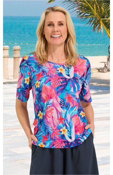 Kurzärmeliges Damenoberteil in pink mit blauem, rosafarbenem und gelbem Blumen- und Blättermuster, rundem Ausschnitt, gerade geschnitten, getragen von lächelnder Frau stehend vor Strandkulisse