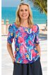 Kurzärmeliges Damenoberteil in pink mit blauem, rosafarbenem und gelbem Blumen- und Blättermuster, rundem Ausschnitt, gerade geschnitten, getragen von lächelnder Frau stehend vor Strandkulisse