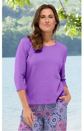 Violetter Pullover mit Strukturmuster, rundem Ausschnitt, dreiviertellangen Ärmeln, gerade geschnitten, getragen von stehender Frau mit Hand in Hosentasche, kombiniert mit bunter Hose mit floralem Muster