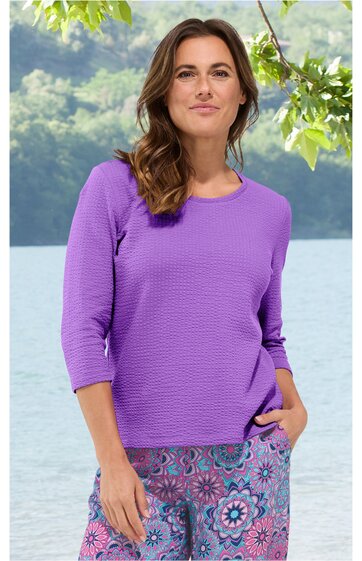 Violetter Pullover mit Strukturmuster, rundem Ausschnitt, dreiviertellangen Ärmeln, gerade geschnitten, getragen von stehender Frau mit Hand in Hosentasche, kombiniert mit bunter Hose mit floralem Muster